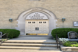 Katholische Kirche St. Anna, ein Gebäude mit einer Tür, Treppe, Geländer, Pflanzen und einer Tafel mit Text darauf, beleuchtet von Lichtern.