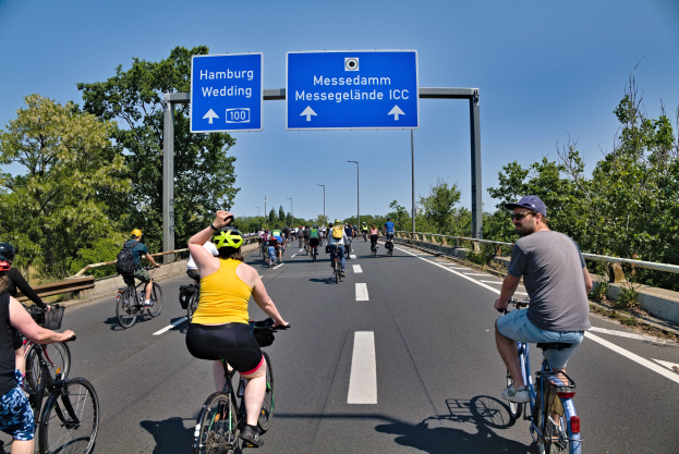 Gruppe von Radfahrern mit Helmen auf einer Straße mit einem Geländer und Bäumen, Laternen und einem klaren blauen Himmel im Hintergrund, mit einer Tafel, die eine Fahrradtour in Hamburg oben anzeigt.