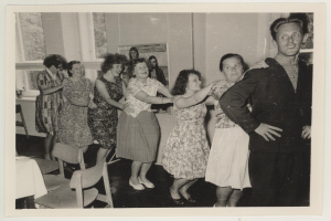 Eine Gruppe von Menschen, die in einem Raum mit Stühlen und einem mit einer wei├čen Decke bedeckten Tisch tanzen. An der Wand befindet sich ein Foto und im Hintergrund sind Fenster zu sehen.
