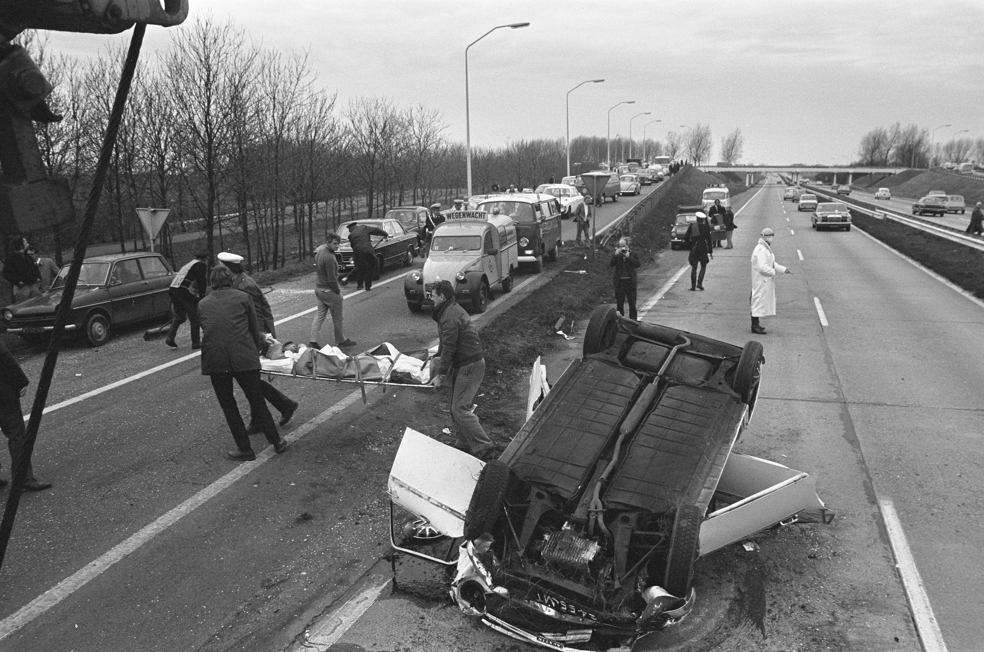 Schwarzes und weißes Bild eines umgestürzten Autos am Straßenrand mit beschädigten Fahrzeugen in der Nähe und einer Gruppe von Menschen drumherum, mit Laternenpfählen, Bäumen, einer Brücke und Himmel im Hintergrund.