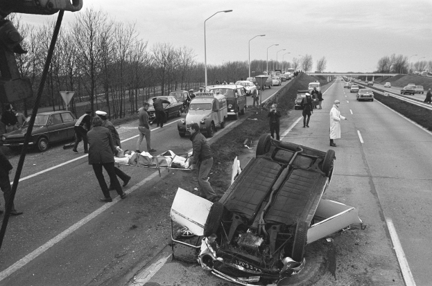 Schwarzes und weißes Bild eines umgestürzten Autos am Straßenrand mit beschädigten Fahrzeugen in der Nähe und einer Gruppe von Menschen drumherum, mit Laternenpfählen, Bäumen, einer Brücke und Himmel im Hintergrund.