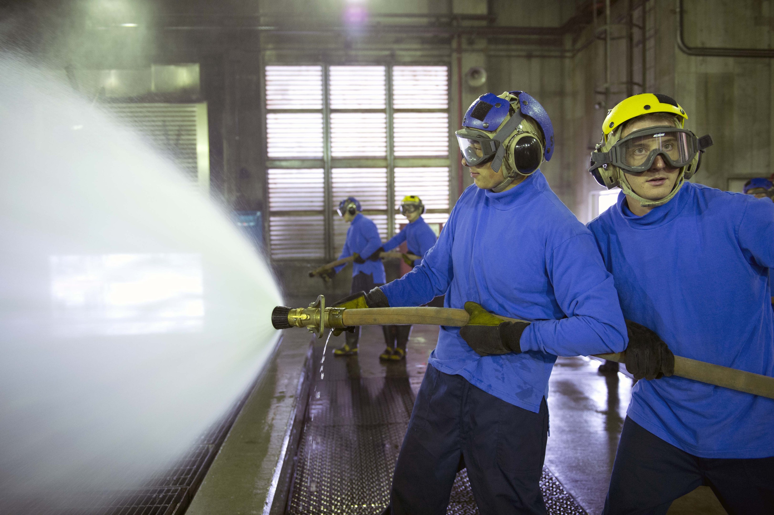 Gruppe von Männern in blauen Hemden und gelben Helmen bei der Arbeit an Maschinen, einer sprüht Wasser auf den Boden in einer Fabrikumgebung mit sichtbaren Rohren, Fenstern und Lichtern.