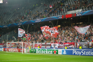 Ein Fussballspiel wird in einem hell erleuchteten Stadion mit einer grossen Zuschauermenge, grünem Rasen, einem Torpfosten mit Bannern und einem Bildschirm in der oberen linken Ecke gespielt.