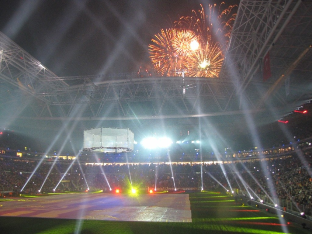 Hell erleuchtetes Olympisches Stadion in London bei Nacht mit grünem Rasen und einer großen Menge Menschen, unter einem Himmel voller Feuerwerk.