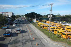 Eine belebte Straße voller Autos, Lastwagen, Busse, Verkehrskegel, Laternenmasten, Strommasten mit Drähten und Schilder mit Text, mit Maschendrahtzaun auf der rechten Seite, Bäume, Gebäude und ein bewölkter Himmel im Hintergrund.