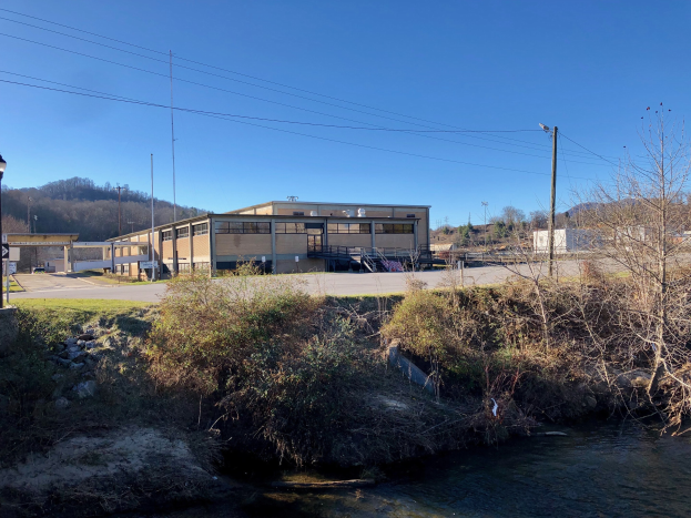 Ein Fluss fließt durch ein grünes Feld neben einem Gebäude, mit Bäumen, Pflanzen, Gras, Steinen, Straßenlaternen, Strommasten, Stromkabeln, Hügeln und einem klaren blauen Himmel im Hintergrund; das Gebäude ist als Standort für eine Wasseraufbereitungsanlage geplant.