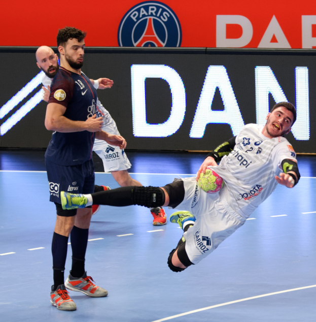Eine Gruppe von Männern, die auf einem Handballfeld spielen, wobei einer den Ball hält und ein Paris-Saint-Germain-Futsal-Siegertafel im Hintergrund zu sehen ist.