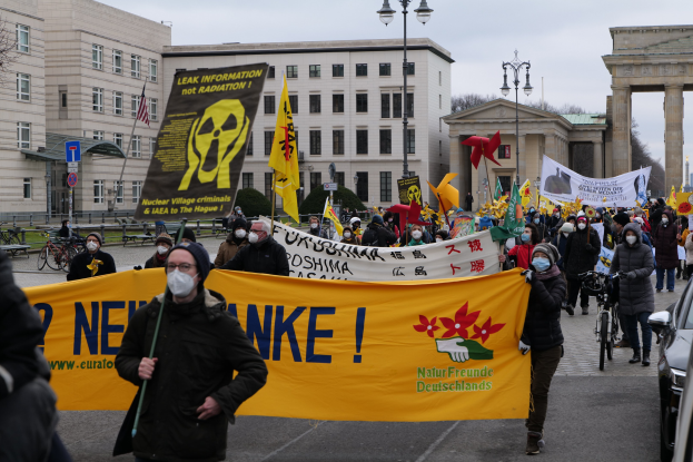 Große Gruppe von Menschen bei einer Straßendemonstration gegen Atomkraft in Deutschland, mit Transparenten und Fahnen, mit Fahrzeugen auf der rechten Seite und Gebäuden, Laternen, Bäumen und einem Tor im Hintergrund.