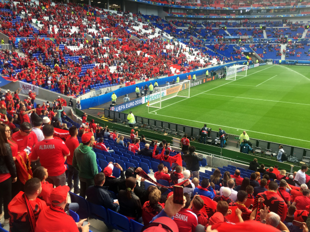 Ein volles Stadion mit Zuschauern, einige sitzen, andere stehen, mit Fahnen, einem Tor und Deckenleuchten, während die Menge gespannt auf den Beginn eines Fußballspiels wartet.