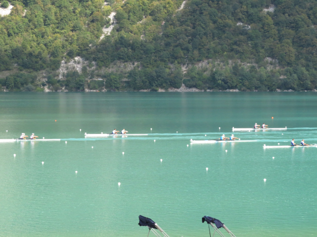 Eine Gruppe von Menschen, die in einem Wettbewerb auf einem Gewässer mit Bäumen und einem Hügel im Hintergrund rudern, mit Kleidung, die über Stühle am Boden gehängt ist.