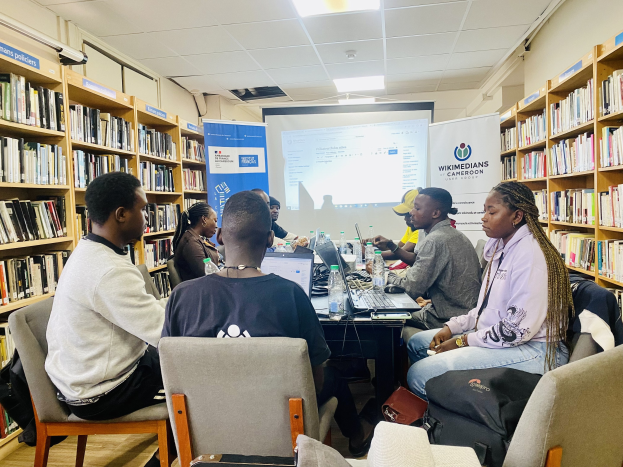 Eine Gruppe von Menschen sitzt um einen Tisch in einer Bibliothek, umgeben von Bücherregalen, in eine Diskussion vertieft mit Laptops, Flaschen und Taschen auf und um den Tisch herum.