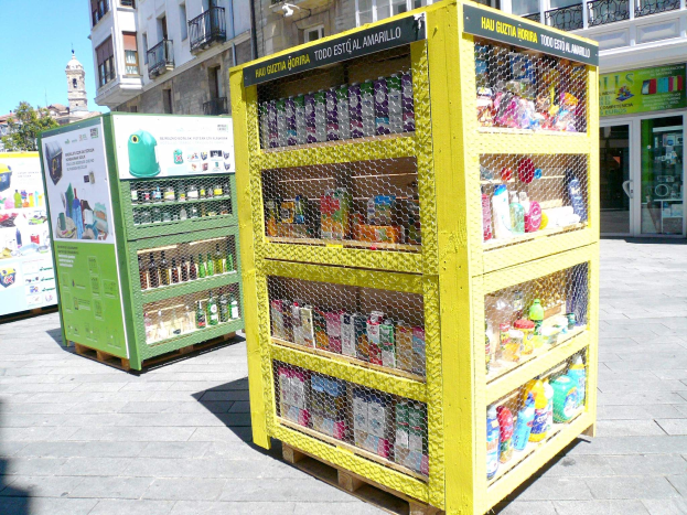 Ein gelber Verkaufsautomat auf einer Stadtstraße, gefüllt mit verschiedenen Gegenständen, umgeben von Gebäuden, Bäumen und einem klaren blauen Himmel.
