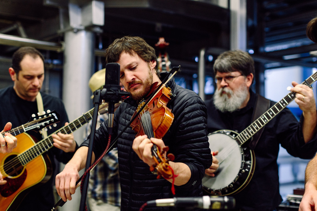 Eine Gruppe von Musikern in schwarzer Kleidung, die verschiedene Instrumente spielen, mit einem Geigenspieler vor einem Mikrofon und einem Gebäude im Hintergrund.