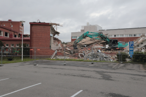 Großes Gebäude in einer Parkanlage mit einem Bagger, Metallstangen, Zaun, Hinweisschild, Pflanzen, Gras, Bäumen, benachbarten Gebäuden und einem bewölkten Himmel.