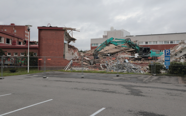 Großes Gebäude in einer Parkanlage mit einem Bagger, Metallstangen, Zaun, Hinweisschild, Pflanzen, Gras, Bäumen, benachbarten Gebäuden und einem bewölkten Himmel.