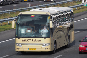 Ein Minibus mit Passagieren fährt auf einer Autobahn neben einem roten Auto, mit Geländern, Gras und texttragenden Säulen im Hintergrund.