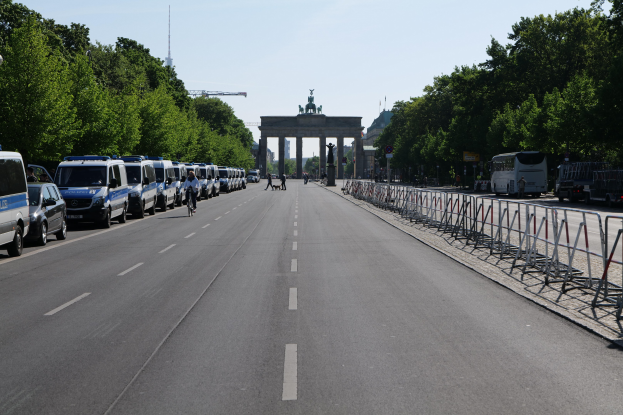 Lange Reihe von Polizeiwagen auf der Seite einer Straße vor dem Brandenburger Tor in Berlin, Deutschland, mit Menschen auf Fahrrädern und in der Nähe Stehenden, Absperrungen, Bäumen, einem Bogen mit Statuen im Hintergrund und sichtbarem Himmel.