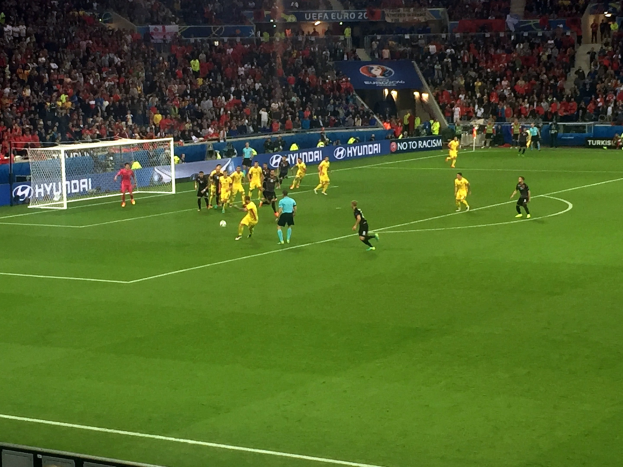 Eine Gruppe von Menschen, die auf einem Feld in einem Stadion Fußball spielen, mit einem Netz auf der linken Seite, Zuschauern auf den Rängen und beleuchteten Tafeln im Hintergrund.