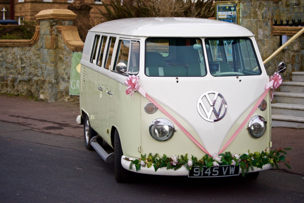 Ein weißer und pinker VW-Campervan mit floralen Verzierungen, der vor einer Steinmauer geparkt ist, neben einer Treppe und einem Gebäude im Hintergrund.