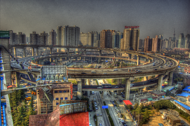 Eine Stadtansicht mit einer zentralen Ringstraße mit Fahrzeugen, einer unteren Straße mit Autos und Fahrrädern, zahlreichen Hochhäusern im Hintergrund, einem Himmel oben, Bäumen auf der linken unteren Seite und kleinen Häusern, die überall in der Stadt verteilt sind.