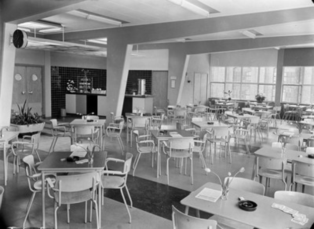 Schwarz-weißes Foto einer Cafeteria mit Tischen und Stühlen, Tellern, Besteck, Gläsern und Blumenvasen auf den Tischen, Türen und Fenstern an der Wand und Deckenlampen.