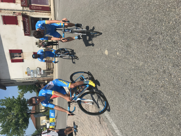 Eine Gruppe von Radfahrern fährt eine Straße entlang an einem Gebäude mit Fenstern und Texttafeln vorbei, Bäume und ein blauer Himmel im Hintergrund, mit Zuschauern auf der Straße darunter.