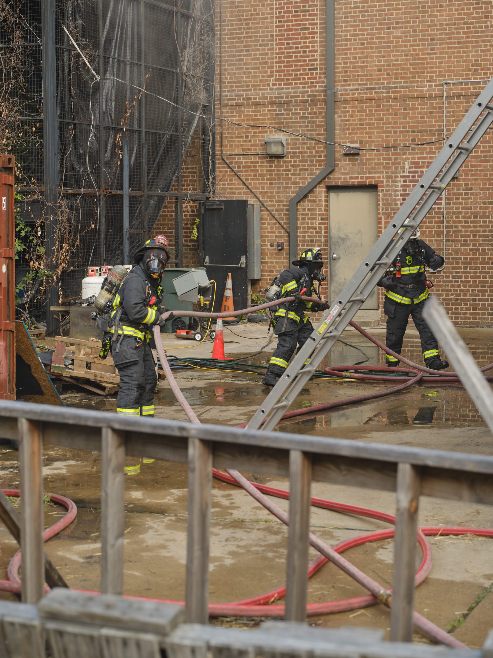 Feuerwehrleute in Helmen arbeiten daran, ein Gebäude Feuer zu löschen, umgeben von Ausrüstung und einem Metallzaun.