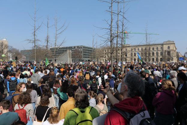 Eine große Menschenmenge steht vor einem Gebäude mit Fenstern, Bäumen und einem klaren Himmel, viele halten Schilder und tragen Taschen, was auf eine Klimaprotest Demonstration hindeutet.
