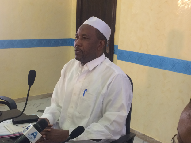 Ein Mann in weißem Anzug und Hut sitzt an einem Tisch mit Mikrofonen, spricht, während Papiere und Gegenstände auf dem Tisch zu sehen sind, und ein weiterer Mensch mit Brille zu seiner Rechten, in einem Raum mit einer Wand und einer Tür im Hintergrund.