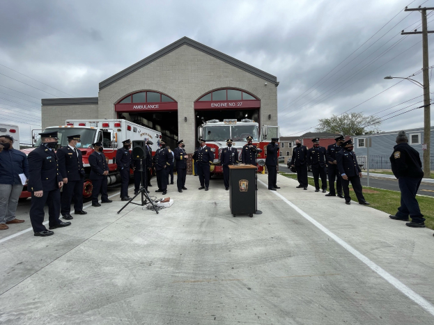 Gruppe von Menschen in Kappen neben einem Löschfahrzeug auf einem Podium während einer Feuerwache-Eröffnung, mit Gebäuden, Bäumen und Strommasten im Hintergrund.