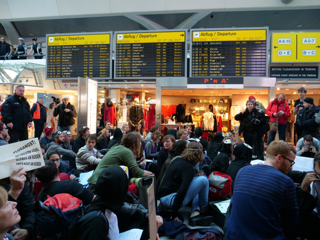 Eine große Gruppe von Menschen in einem Flughafen, einige mit Taschen und Papieren sitzend, andere stehend, mit Texttafeln, Schaufensterpuppen in Kleidern und Deckenlampen im Hintergrund, was auf eine Demonstration hindeutet.