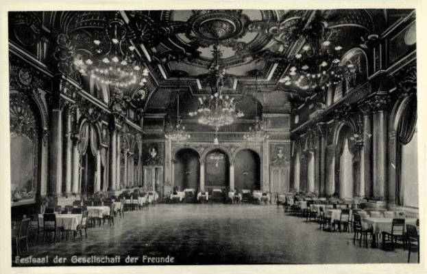 Schwarzes und weißes Foto eines großen Raums mit Tischen, Stühlen, Säulen, Bögen und Kronleuchtern am Himmel, beschriftet mit "Fest der Gesellschaft der Freunde, Berlin" unten.