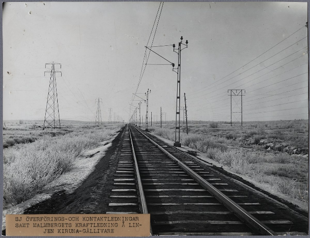 Schwarzes und weißes Foto einer Schienenstrecke mit elektrischen Masten und Drähten daneben, umgeben von Gras auf beiden Seiten, mit Text unten.