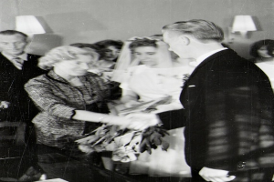 Ein Schwarz-Weiß-Foto eines Brautpaares beim Austausch der Eheringe, wobei die Braut ein weißes Kleid und einen Schleier trägt und einen Blumenstrauß hält, und der Bräutigam einen Anzug und eine Krawatte trägt, mit Wandlampen im Hintergrund.