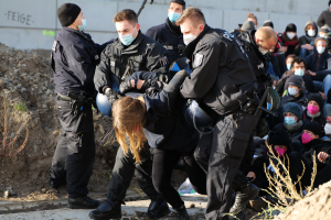 Gruppe maskierter Personen mit Mützen, einige mit Waffen, umringt eine Frau, die von der Polizei eskortiert wird, in einer städtischen Umgebung mit Pflanzen, Steinen und einer Wand mit protestrelevanten Texten.