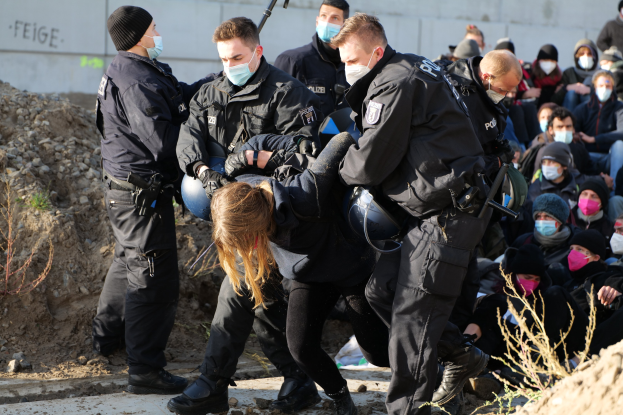 Gruppe maskierter Personen mit Mützen, einige mit Waffen, umringt eine Frau, die von der Polizei eskortiert wird, in einer städtischen Umgebung mit Pflanzen, Steinen und einer Wand mit protestrelevanten Texten.
