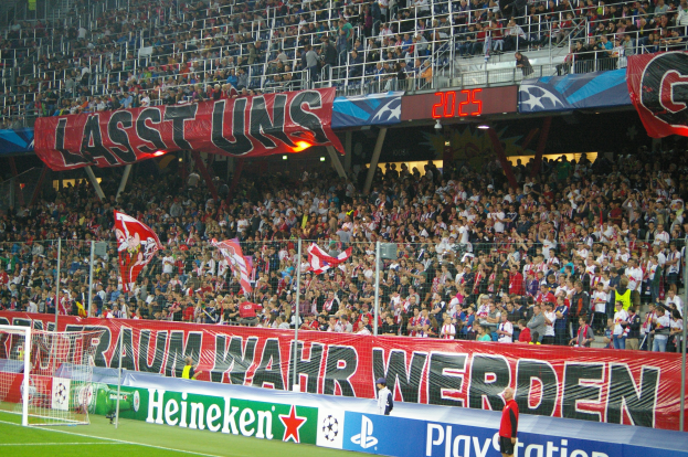 Ein Fußballspiel in einem Stadion mit einer großen Zuschauermenge, einem saftig grünen Rasen, einem Torpfosten links, Bodenbanner und Menschen mit Fahnen in der Nähe, mit Geländern, Tafeln und Lampen im Hintergrund.