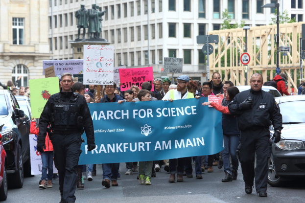 Gruppe von Menschen marschiert mit einem "March for Science Frankfurt am Main"-Schild die Straße entlang, Autos und Gebäude im Hintergrund.