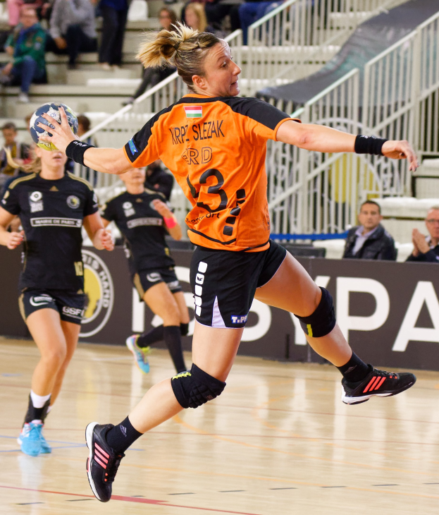Eine Frau in einem orangen T-Shirt und schwarzen Shorts hält einen Handball, mit Zuschauern auf Treppen und Geländern im Hintergrund hinter einem Banner mit der Aufschrift "Krp Slezak - Handball-Weltmeisterschaft 2019."