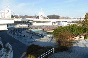 Blick auf das Olympische Stadion von einem hohen Standpunkt aus, mit verschiedenen Objekten und Grünflächen im Vordergrund und einem bewölkten Himmel im Hintergrund.