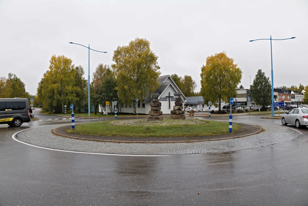 Ein Kreisverkehr mit parkenden Autos, einem Van und einem Auto im Vordergrund, umgeben von Straßenlaternen, Bäumen, Gebäuden und dem Himmel im Hintergrund.