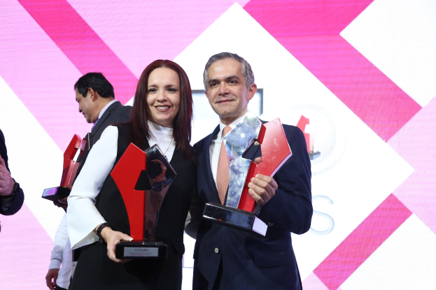 Zwei strahlende Individuen auf einer Bühne mit Trophäen, mit einem Bildschirm im Hintergrund.