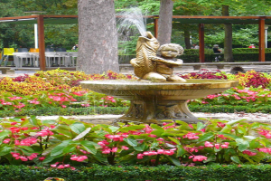 Ein Brunnen thront in der Mitte eines grünen Parks, umgeben von Pflanzen und Blumen, mit Bäumen, Menschen, Bänken und einer Hütte im Hintergrund.