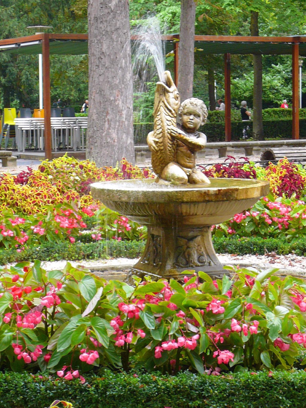 Ein Brunnen thront in der Mitte eines grünen Parks, umgeben von Pflanzen und Blumen, mit Bäumen, Menschen, Bänken und einer Hütte im Hintergrund.