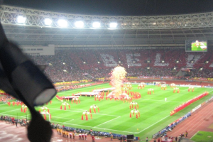 Ein Stadion mit Flutlicht, Eisengittern, Zuschauern, stehenden Personen, Werbetafeln, Anzeigetafeln, Überwachungskamera, Flaggen und Dekorationselementen.