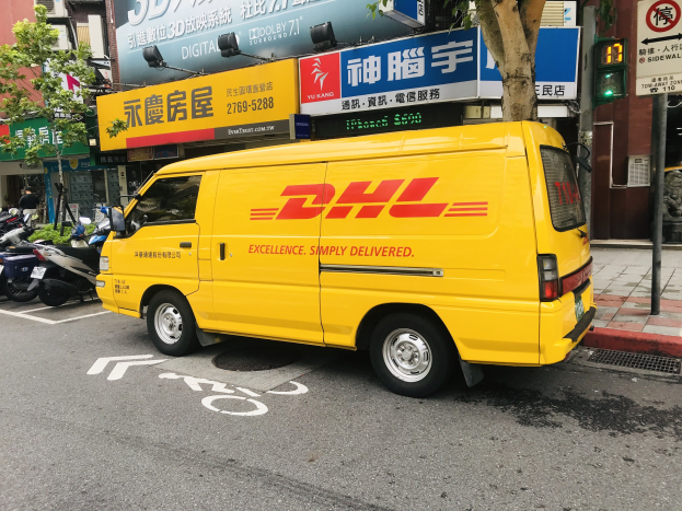 Gelbes DHL-Lieferfahrzeug auf der Straße geparkt, umgeben von Gebäuden, Bäumen, Schildern, Pfählen, Verkehrssignalen und einer Person auf dem Gehweg.