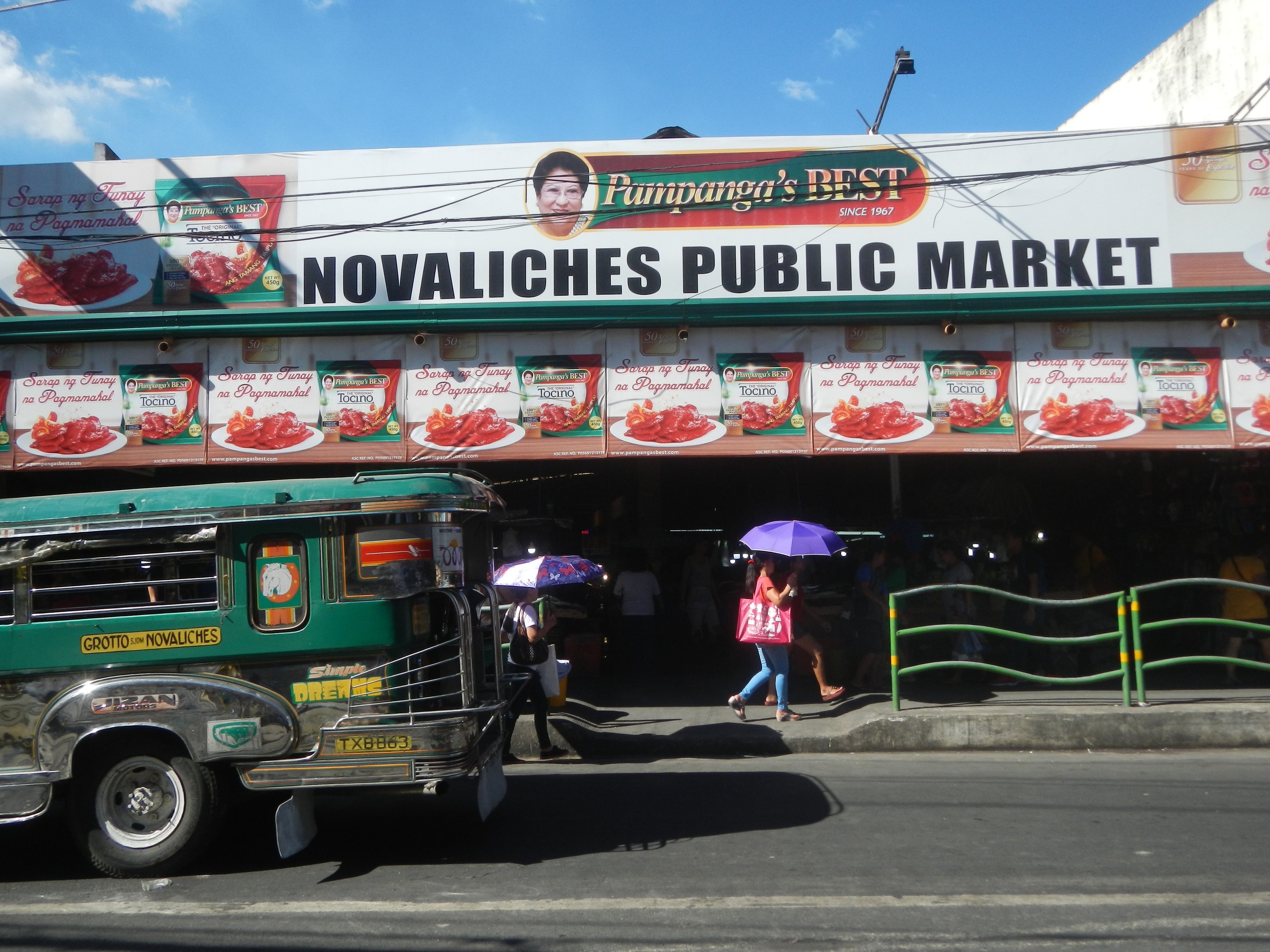 Ein Bus fährt an dem Gebäude des Novaliches Public Market vorbei, während Fußgänger Regenschirme halten, eine Begrenzung auf der rechten Seite und Gebäude, ein Lichtmast und Drähte im Hintergrund.