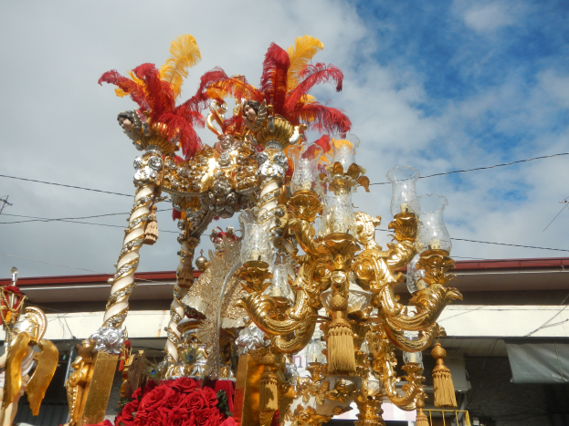 Ein großes goldenes und rotes Festwagen mit Blumen und anderen Schmuckelementen geschmückt in einem Karnevalsumzug, mit einem Gebäude, Strommasten mit Drähten und einem bewölkten Himmel im Hintergrund.