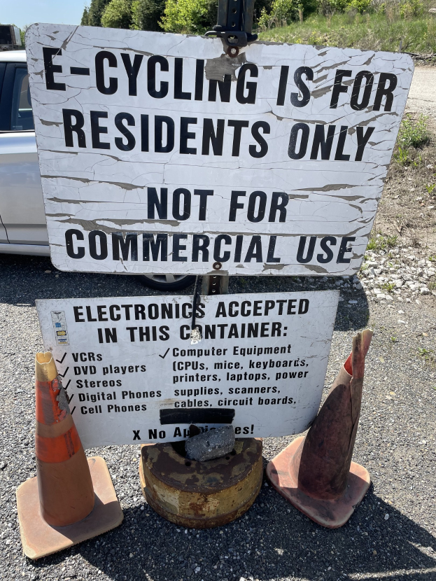 Schild mit der Aufschrift "E-Bike-Fahren nur für Anwohner" neben einer Straße mit Absperrbaken, einem Fahrzeug, Bäumen und einem klaren blauen Himmel im Hintergrund.