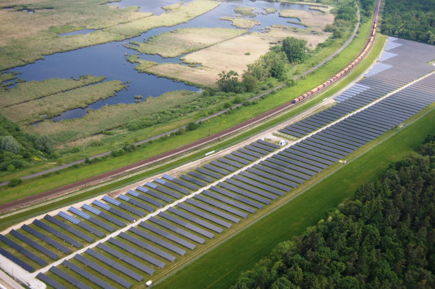Luftaufnahme eines Solarparks mit Solarpanelen auf einem Feld, umgeben von Bäumen, Gras, Pflanzen und Wasser, mit einem Zug auf einem nahen Bahnsteig.